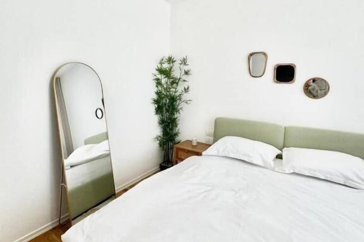 A minimalist bedroom with a bed, a mirror, a plant, and a wall-mounted light fixture.
