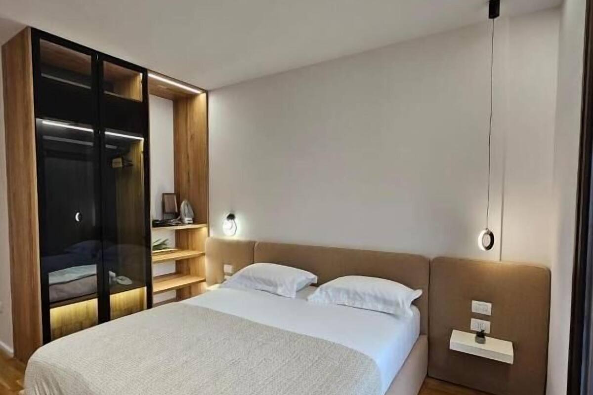 A modern bedroom with a large bed, built-in wardrobe, bedside tables, and a view through a window.