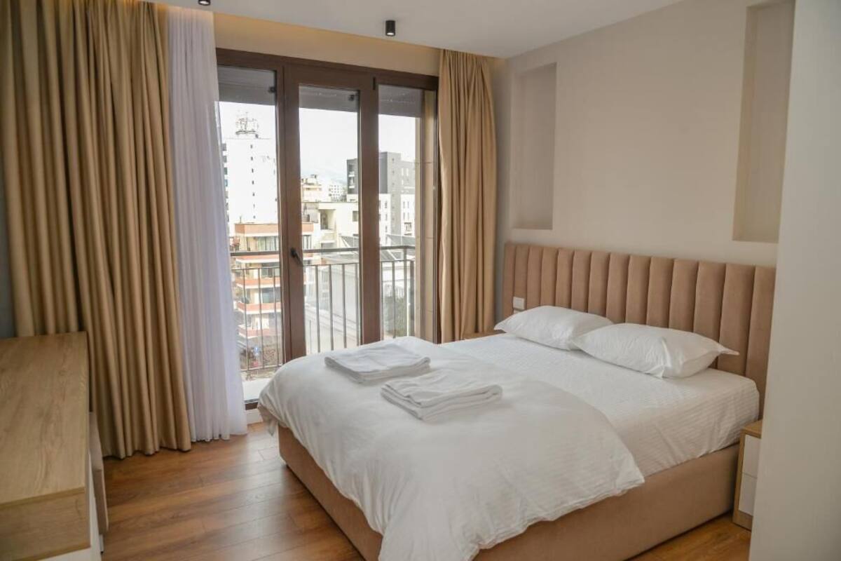 A hotel room with a bed, a wooden desk, and a view of buildings through a glass door.