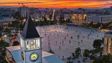 Una plaza con una torre de reloj, rodeada de edificios y un atardecer de fondo.