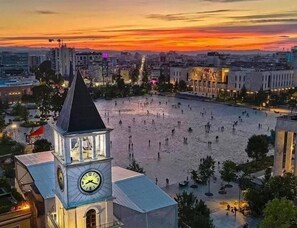 A square with a clock tower, surrounded by buildings and a sunset in the background. - Green Terrace Apartments Tirana (Tirana)