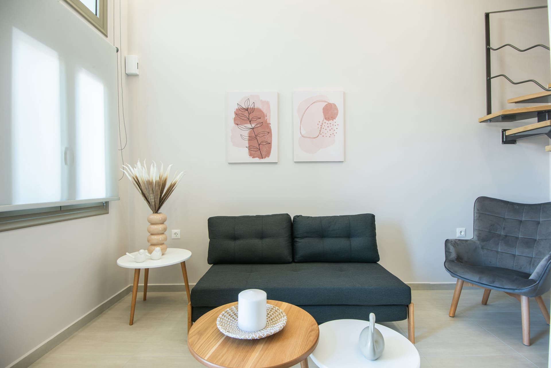 Una sala moderna con un sofá verde oscuro, un sillón gris, una pequeña mesa redonda de madera con una vela blanca y dos cuadros abstractos en la pared.