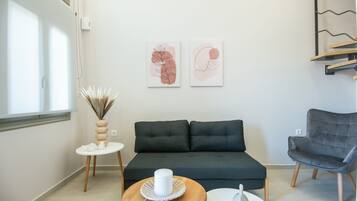 Una sala moderna con un sofá verde oscuro, un sillón gris, una pequeña mesa redonda de madera con una vela blanca y dos cuadros abstractos en la pared.