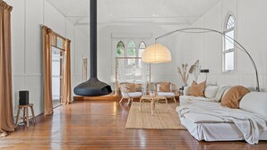 Una habitación amplia con una gran chimenea negra, paredes blancas, suelo de madera, un sofá blanco y un área de comedor con sillas de mimbre y una mesa.
