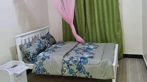 Una cama con una colcha estampada con motivos florales, una mesita de noche blanca con papeles y cortinas verdes.
