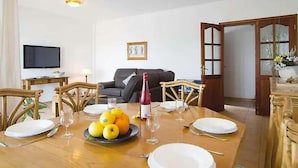 Uma área de refeições com uma mesa de madeira posta para uma refeição, um sofá ao fundo e uma televisão na parede.