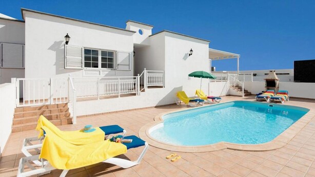 Uma área de piscina com espreguiçadeiras e guarda-sóis, uma escadaria que leva a um deque coberto e um prédio branco com persianas.