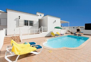 A swimming pool area with lounge chairs and umbrellas, a staircase leading to a covered deck, and a white building with shutters. - Villa Diama With 3 Bedrooms and 2 Bathrooms (Yaiza)