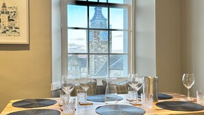 Une table à manger dressée avec des verres à vin et des assiettes, une vue sur un clocher par la fenêtre et un tableau encadré au mur.