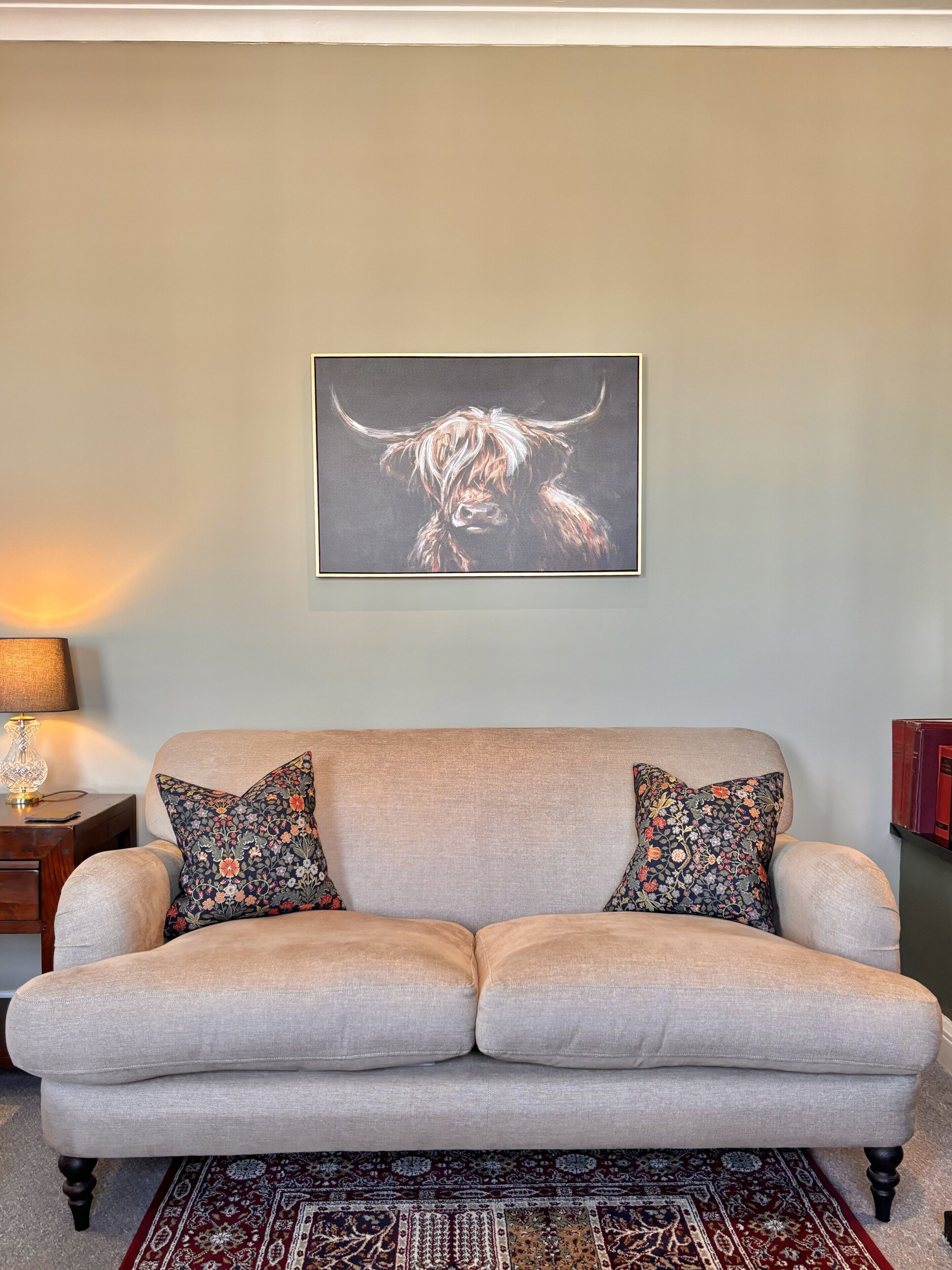 Un salon avec un canapé beige, des coussins à motifs floraux, un tapis à dessins et, au mur, une œuvre d’art encadrée représentant un taureau.