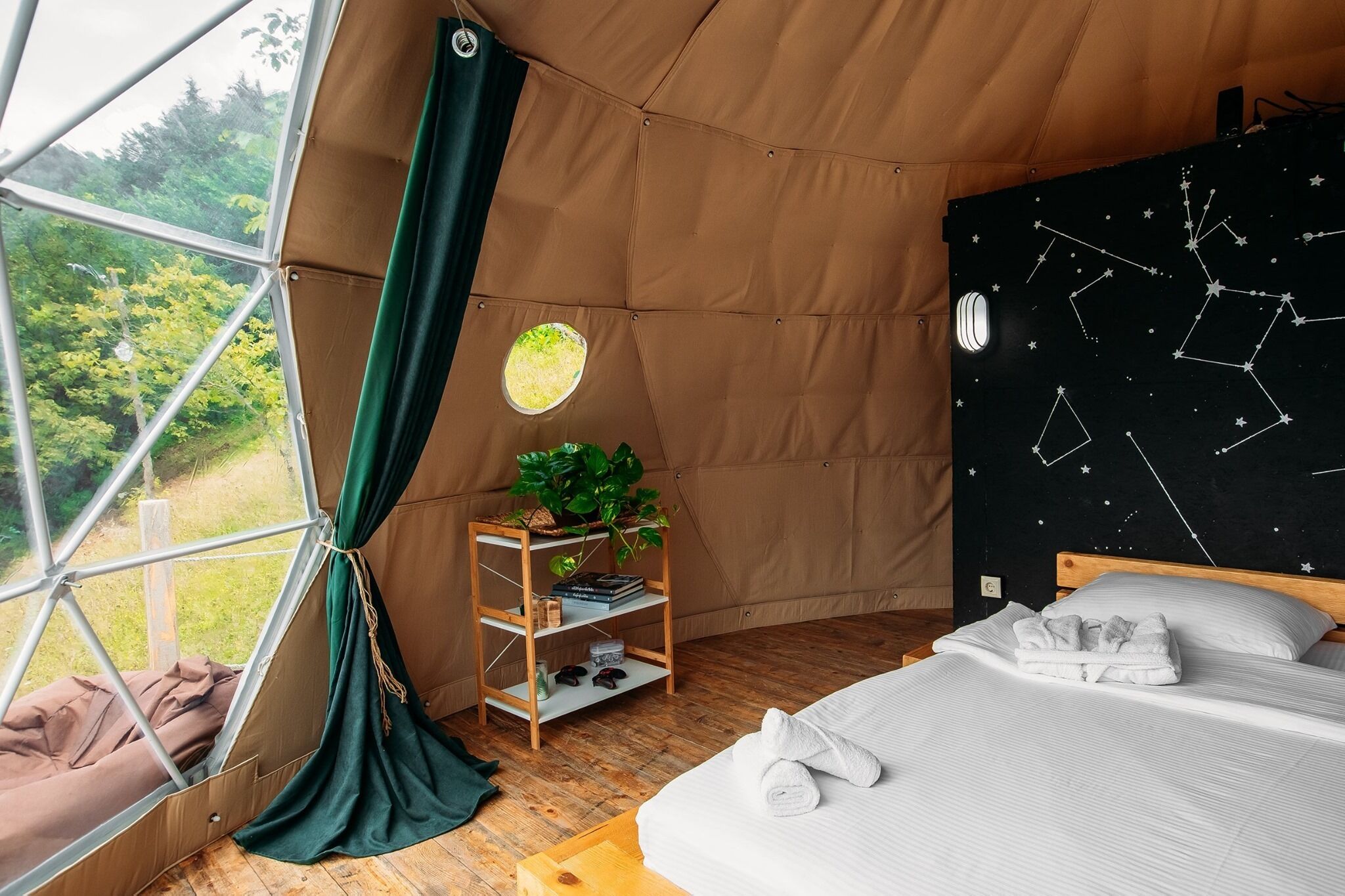 Una habitación tipo tienda de campaña acogedora, con cama, una estantería con plantas y una ventana con cortina.