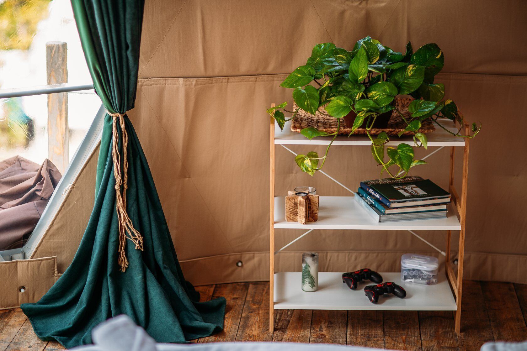 Una habitación acogedora con suelo de madera, cortina verde, estantería con libros y una planta, y una consola de videojuegos.
