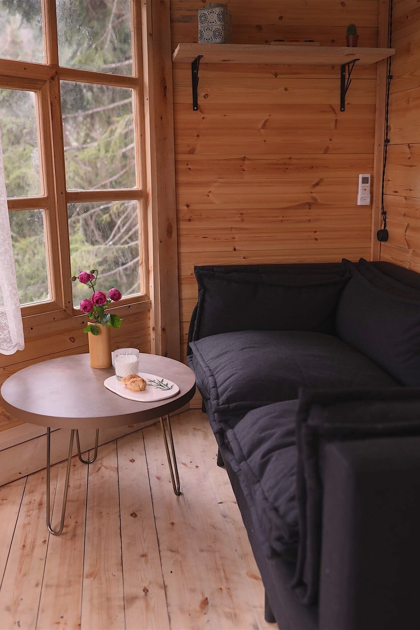 A cozy cabin interior with a wooden wall, a round table with a vase of flowers, a plate with a pastry, and a dark-colored sofa.