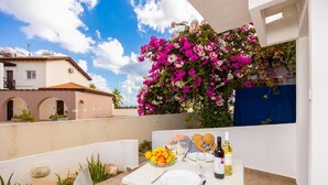 Ein Tisch ist gedeckt mit Tellern, Besteck, einer Obstschale und Weinflaschen auf einer Terrasse mit Blick auf ein Gebäude und einen blühenden Strauch.
