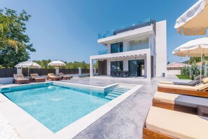 A modern two-story house with a swimming pool, outdoor seating, and umbrellas.
