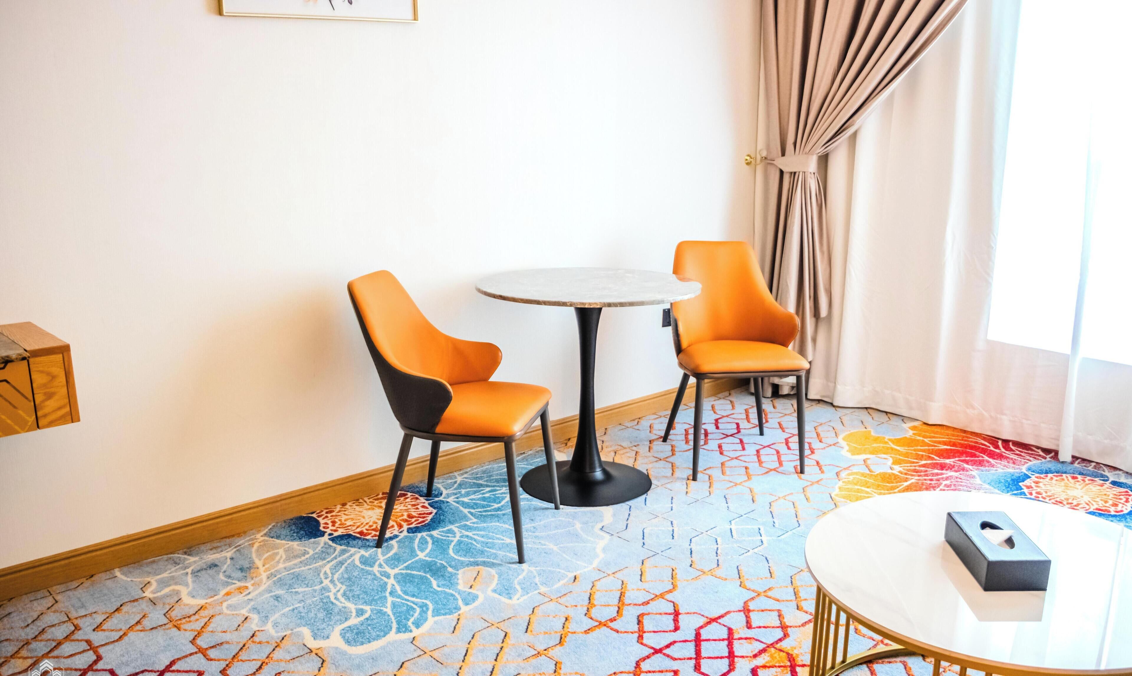 A hotel room with two orange chairs, a small round table, a patterned carpet, and a window with curtains.
