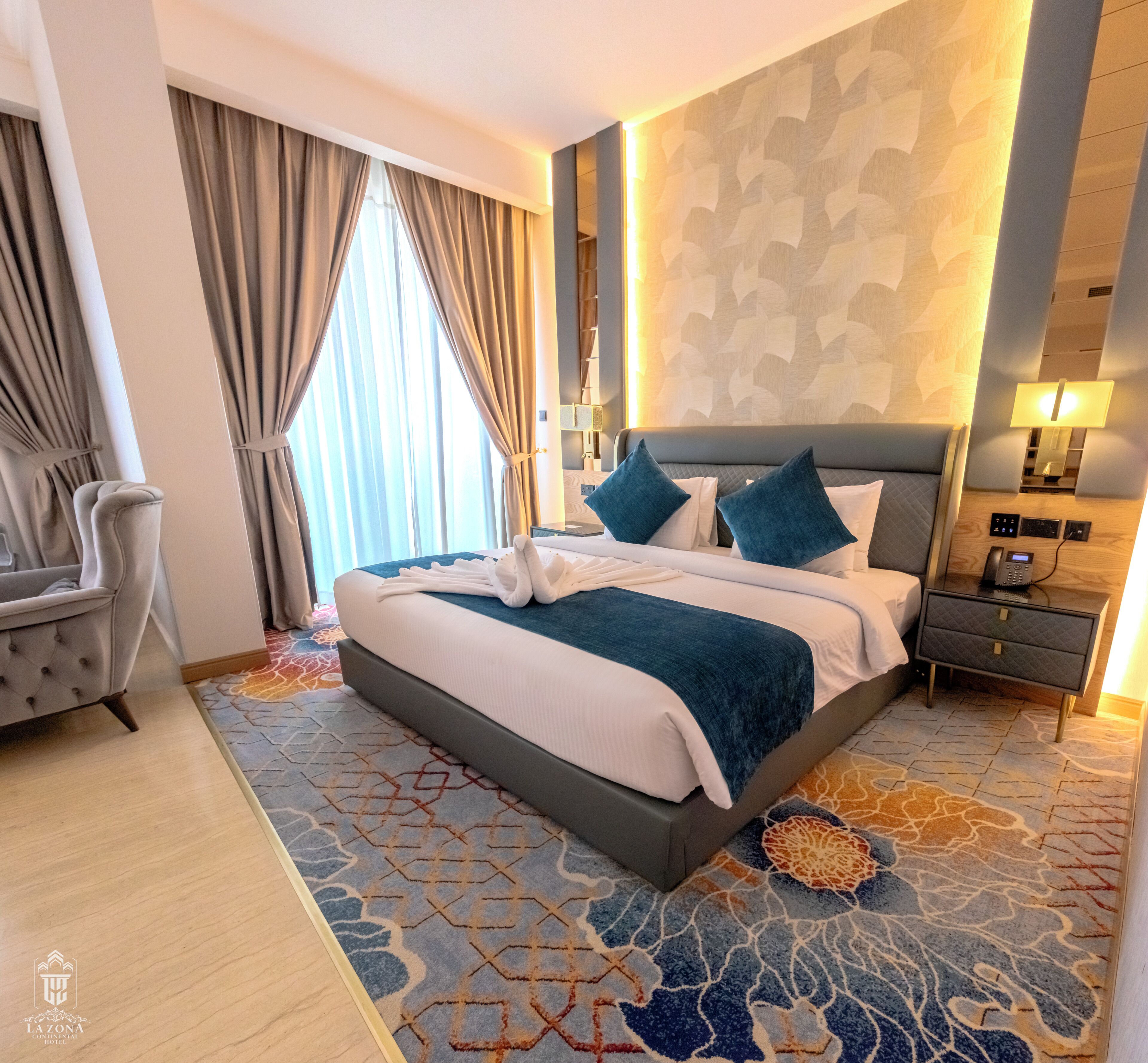 Quarto de hotel com uma cama grande, mesa de cabeceira com abajur, uma cadeira e um tapete estampado.