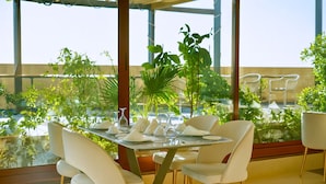 A dining area with a table set for six, surrounded by white chairs with gold legs, and large windows offering a view of greenery and a building. - La Zona Continental Hotel (Dubai)