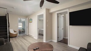 Un salon spacieux doté d’un ventilateur de plafond, d’une table basse ronde en bois et d’un téléviseur à écran plat fixé au mur.
