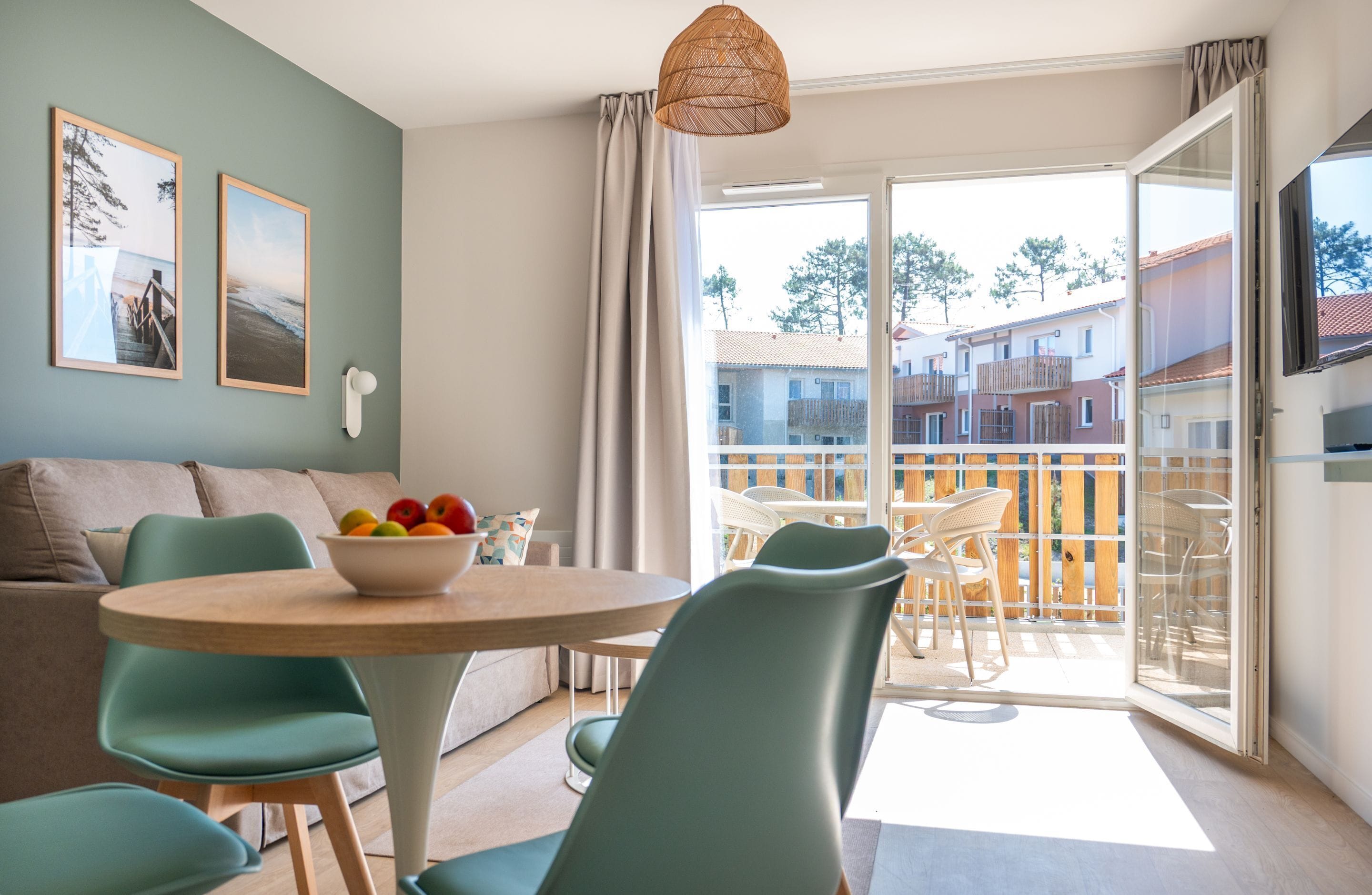 Una sala de estar moderna con una mesa redonda de madera, sillas color turquesa, un sofá y un balcón con mobiliario exterior.