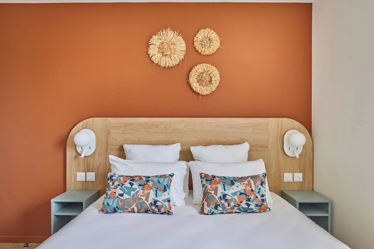 Een netjes opgemaakt bed met witte beddengoed en twee decoratieve kussens, een houten hoofdbord en drie wandlampen.