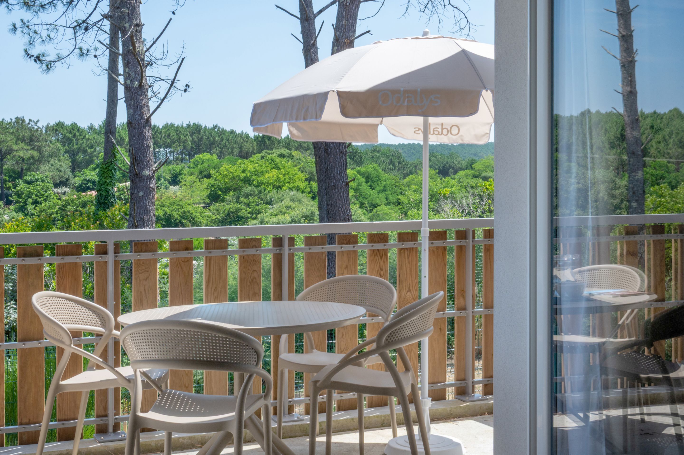 Ein Balkon mit Tisch und Stühlen, einem großen Sonnenschirm und Blick auf Bäume und Grünflächen.
