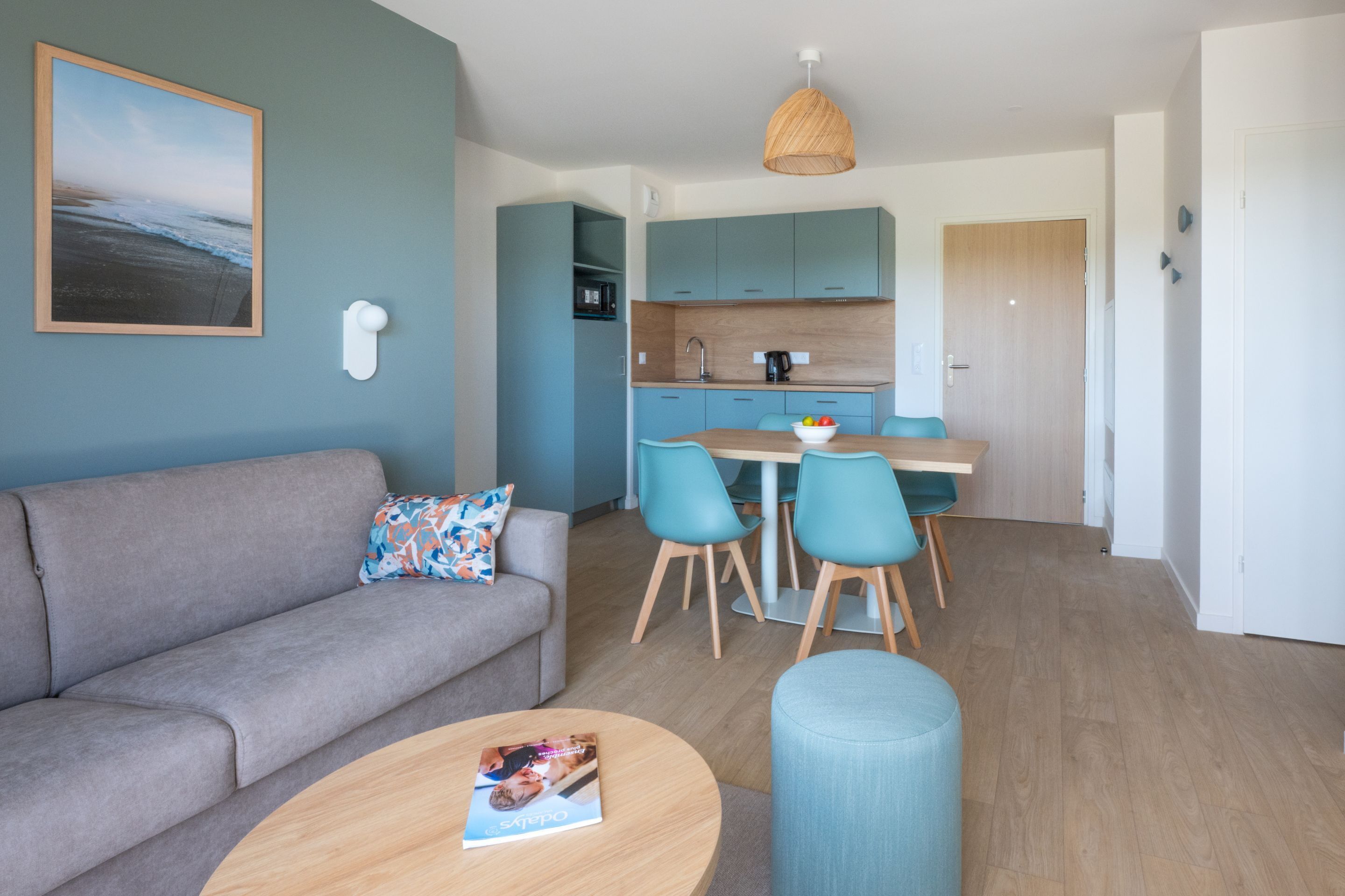 A modern living room with a grey sofa, a round coffee table with a magazine, and a dining area with two chairs and a wooden table.