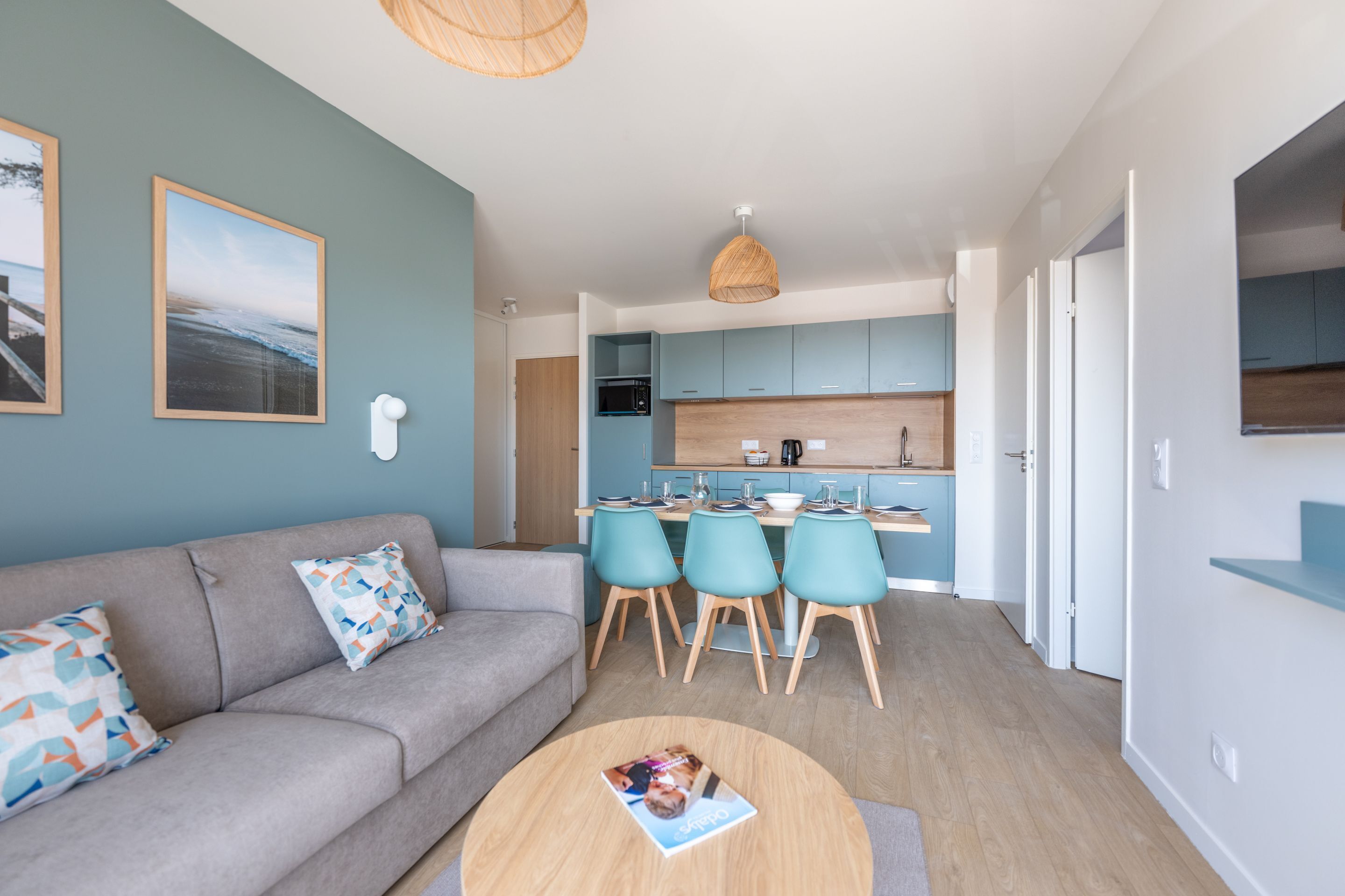 Ein modernes Wohnzimmer mit grauem Sofa, einem Couchtisch und einem Essbereich mit blaugrünen Stühlen und einem Holztisch.