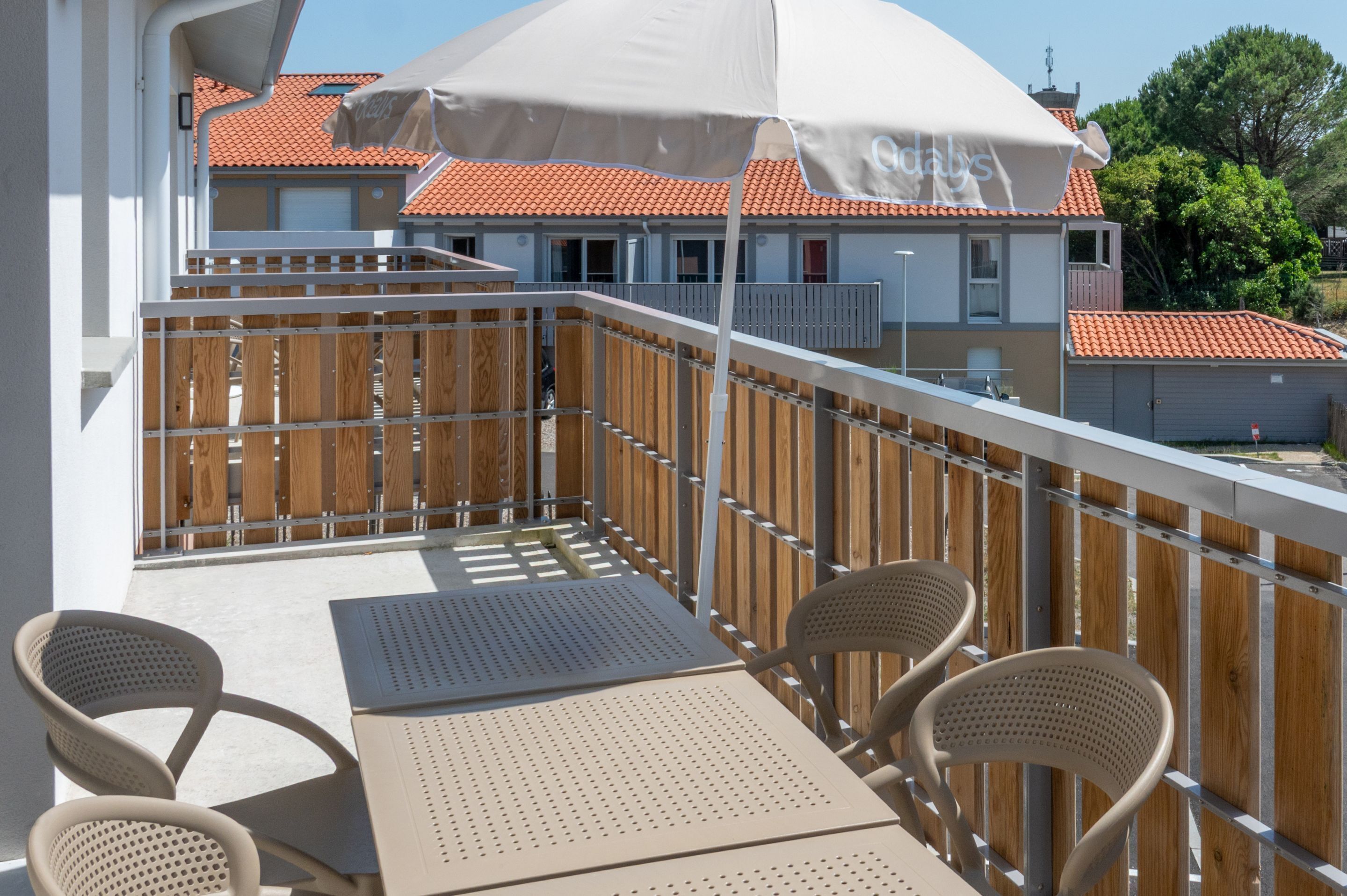 Un balcone con tavolo e sedie, una recinzione di legno e un edificio sullo sfondo con tetto in tegole rosse.