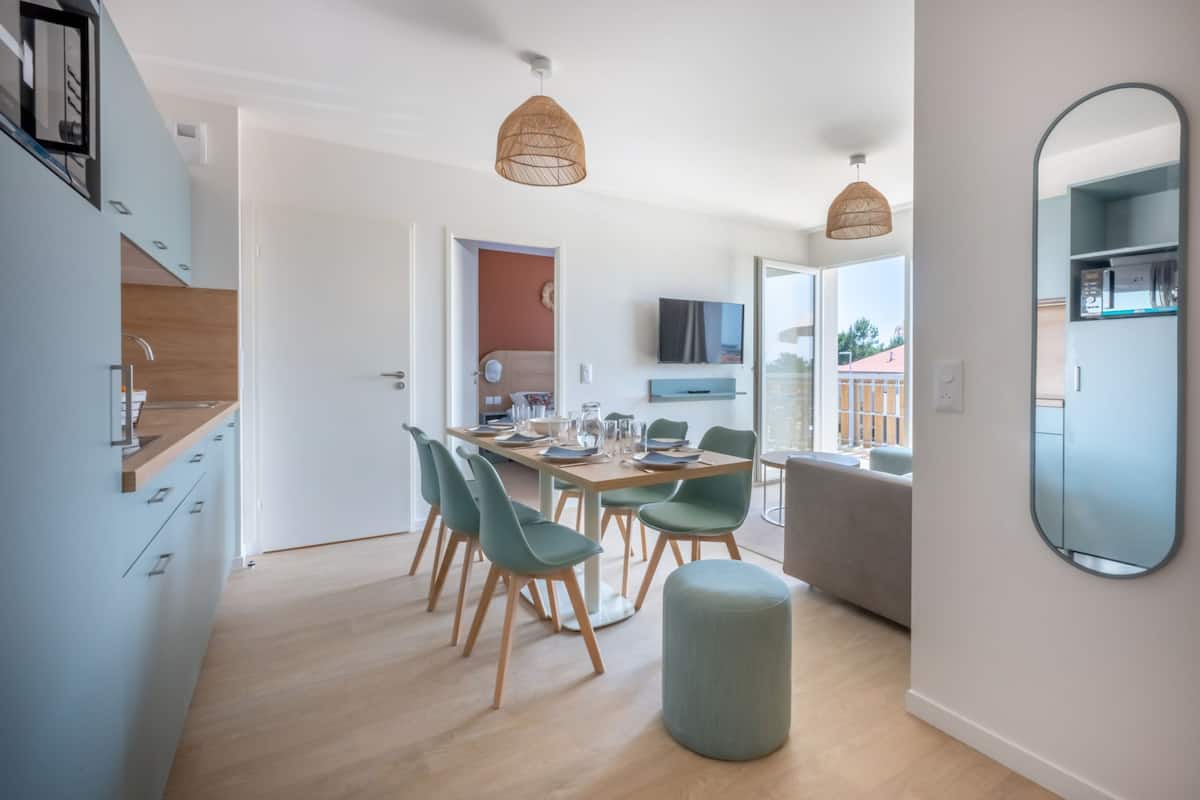 Een moderne keuken met houten kastjes, een eethoek met een tafel en stoelen, en een grote spiegel aan de muur.