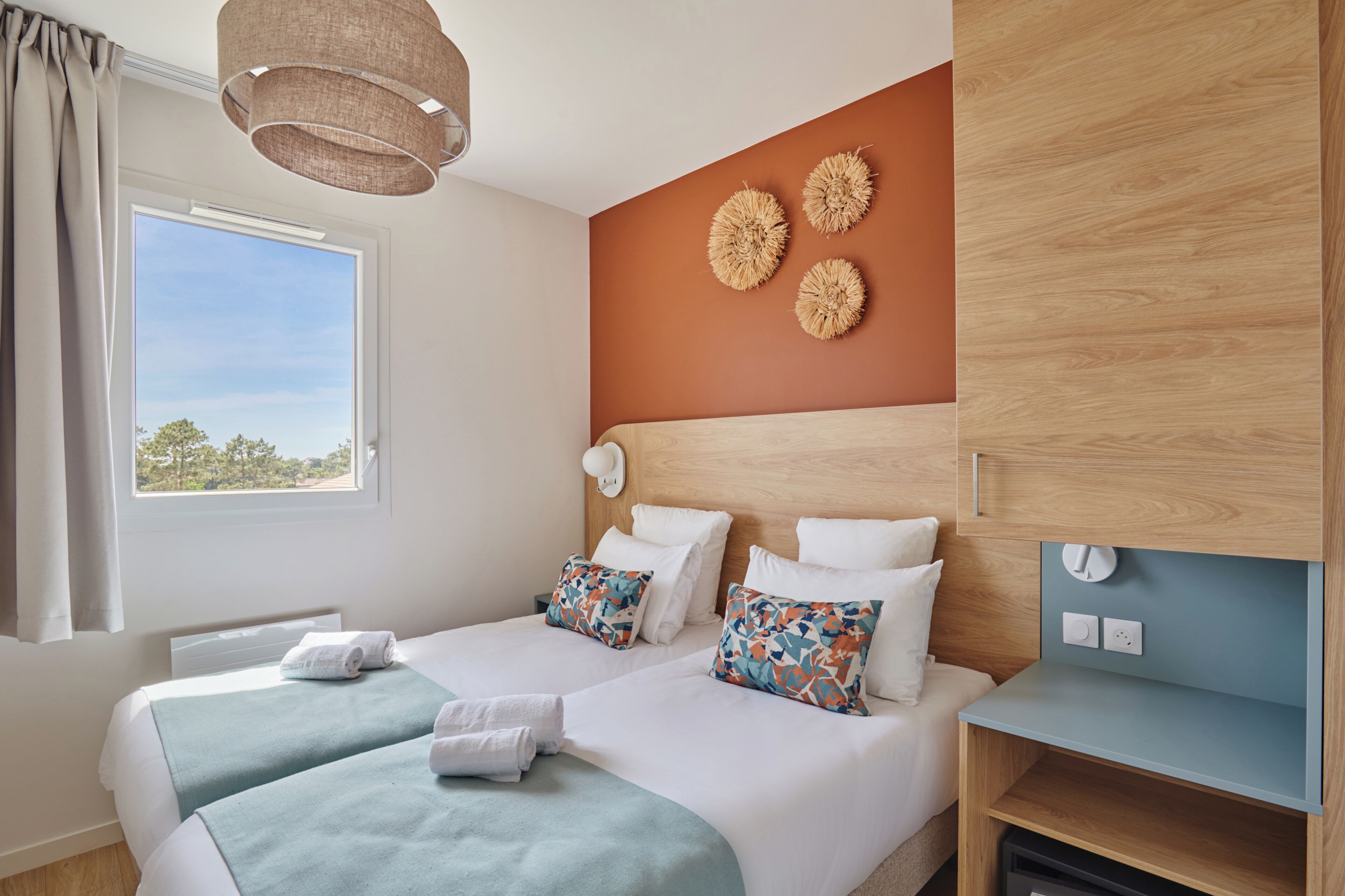 A hotel room with two beds, a window with curtains, a wall-mounted lamp, and a wooden headboard.