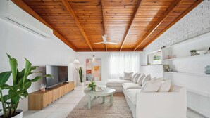 Sala de estar moderna com teto de madeira, parede de tijolos brancos, sofá branco, televisão e planta em vaso.