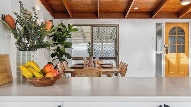 Teto de madeira, uma mesa de jantar com cadeiras, uma fruteira e uma janela com design treliçado.