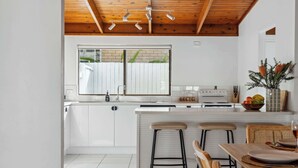 Uma cozinha moderna com armários brancos, teto de madeira, uma janela grande e um espaço de refeições com banquetas.