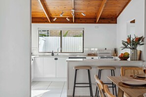 A modern kitchen with white cabinets, a wooden ceiling, a large window, and a dining area with stools.