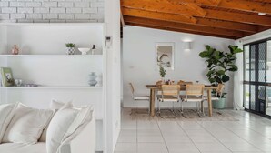 Interior moderno com parede de tijolos brancos, vigas de madeira no teto, área de refeições com mesa e cadeiras, e uma grande planta em vaso.