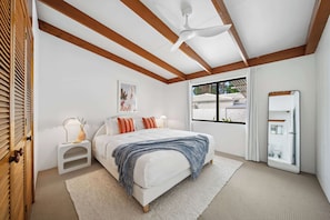 Ein Schlafzimmer mit einem Bett, Nachttischen, einem Deckenventilator, einem Fenster mit Vorhängen und einem Spiegel.