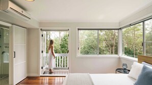 A bedroom with a large bed, a window with a view of trees, a door leading to a balcony, and a small bedside table with a lamp. - Bangalow Retreat in Buderim (Mooloolaba Region)