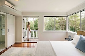 Une chambre avec un grand lit, une fenêtre donnant sur des arbres, une porte menant à un balcon et une petite table de chevet avec une lampe.