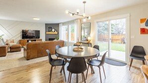 A modern living room with a fireplace, a dining area with a round table, and a large window offering a view of the backyard. - Maple Suite Retreat: Arcade • Ping Pong • HomeGym • Toys • Pool • Playgrounds  (Meridian)
