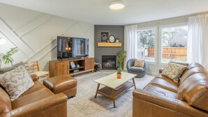 A living room with leather sofas, a coffee table, a TV on a wooden stand, a fireplace, and a large window with curtains. - Maple Suite Retreat: Arcade • Ping Pong • HomeGym • Toys • Pool • Playgrounds  (Meridian)