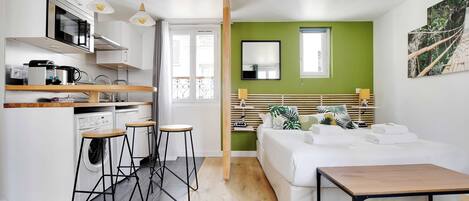 A compact hotel room with a kitchenette, a bed with a wooden headboard, a small table, and a large window.