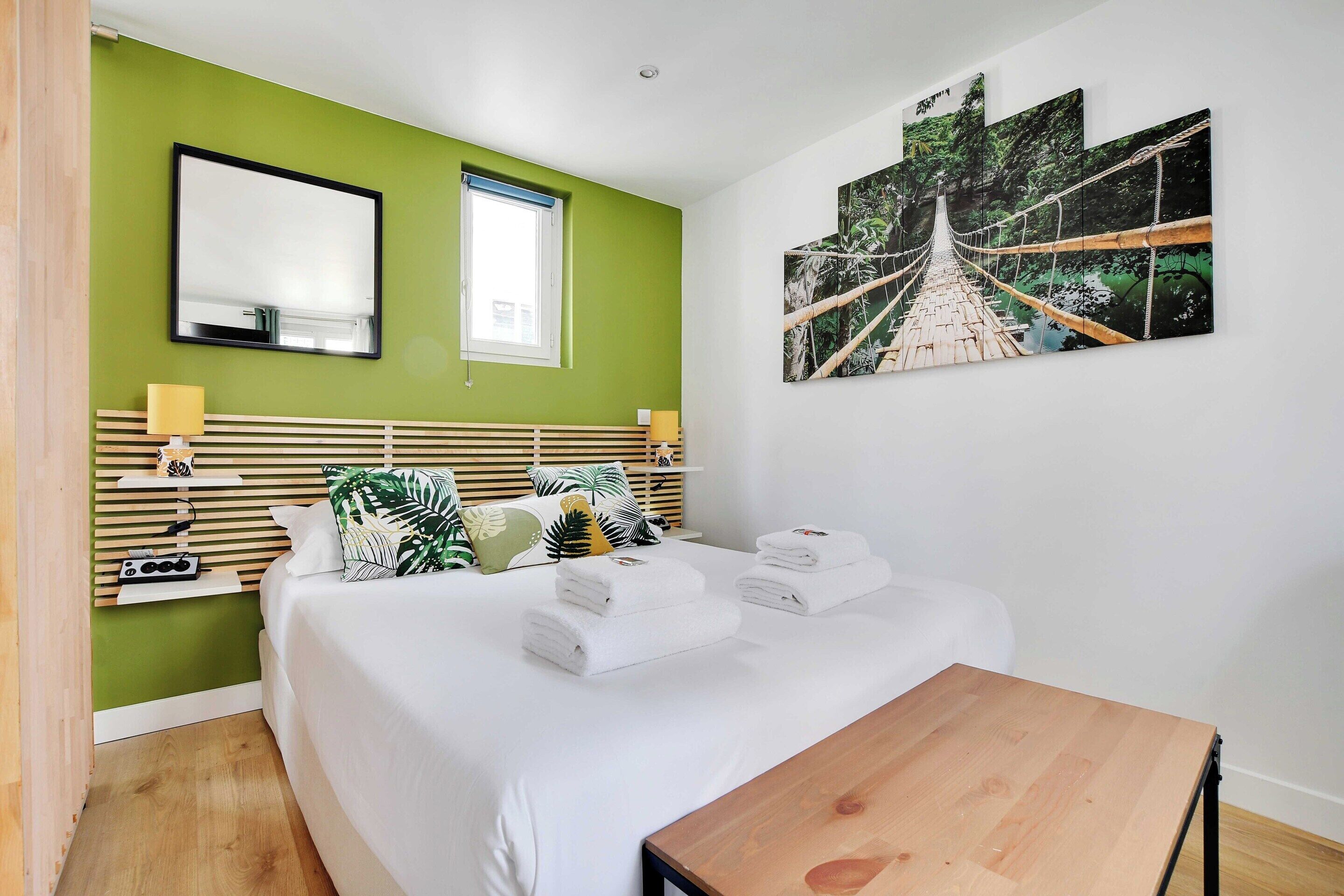 A bedroom with a bed, a wooden headboard, a nightstand, a table, a wall-mounted mirror, a window, and a large painting of a bridge.