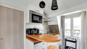 Une cuisine compacte équipée d’un four à micro-ondes, d’une machine à café et d’un petit évier. On y trouve une table en bois et deux tabourets. Une machine à laver est visible dans un coin.