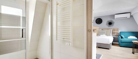 A modern hotel room with a glass shower enclosure, a built-in towel rack, a bed with a wooden headboard, a blue sofa, and an air conditioning unit on the wall.