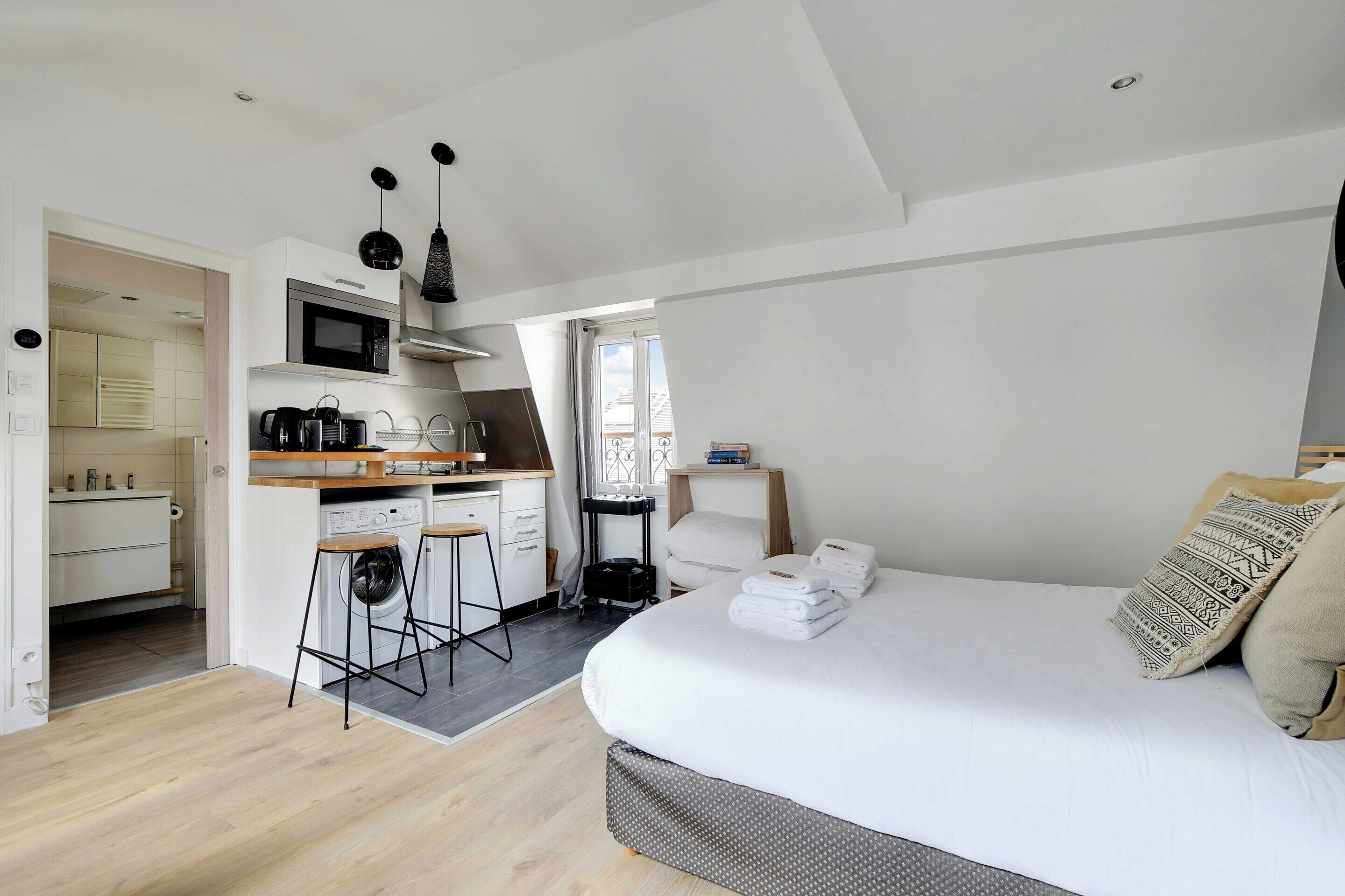 A modern bedroom with a large bed, a small kitchenette, and a bathroom visible through an open door.