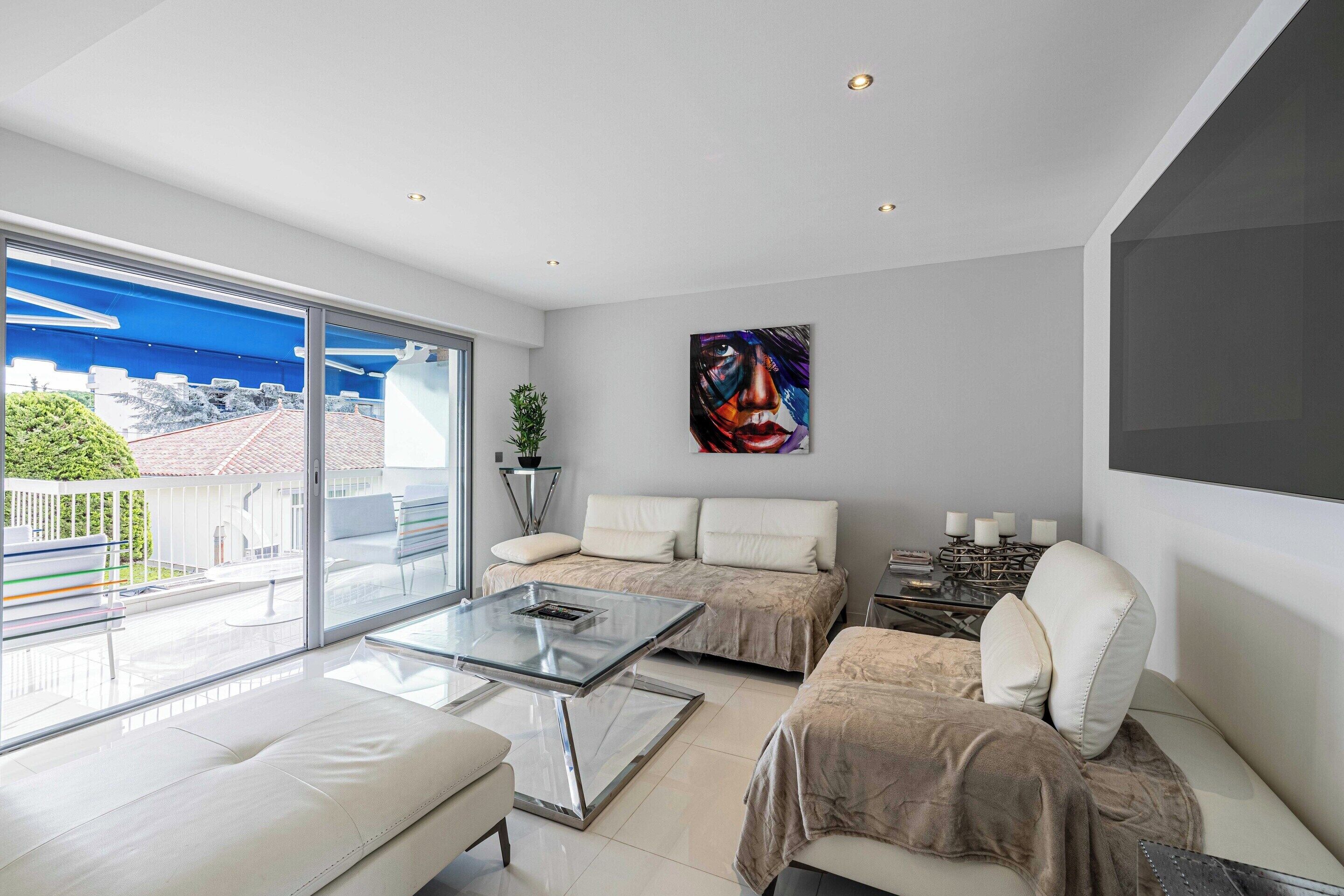 Ein modernes Wohnzimmer mit Glastisch, einer Couch und einem Sessel. An der Wand hängt ein Gemälde und vom Balkon aus hat man Blick auf Gebäude und Grünflächen.