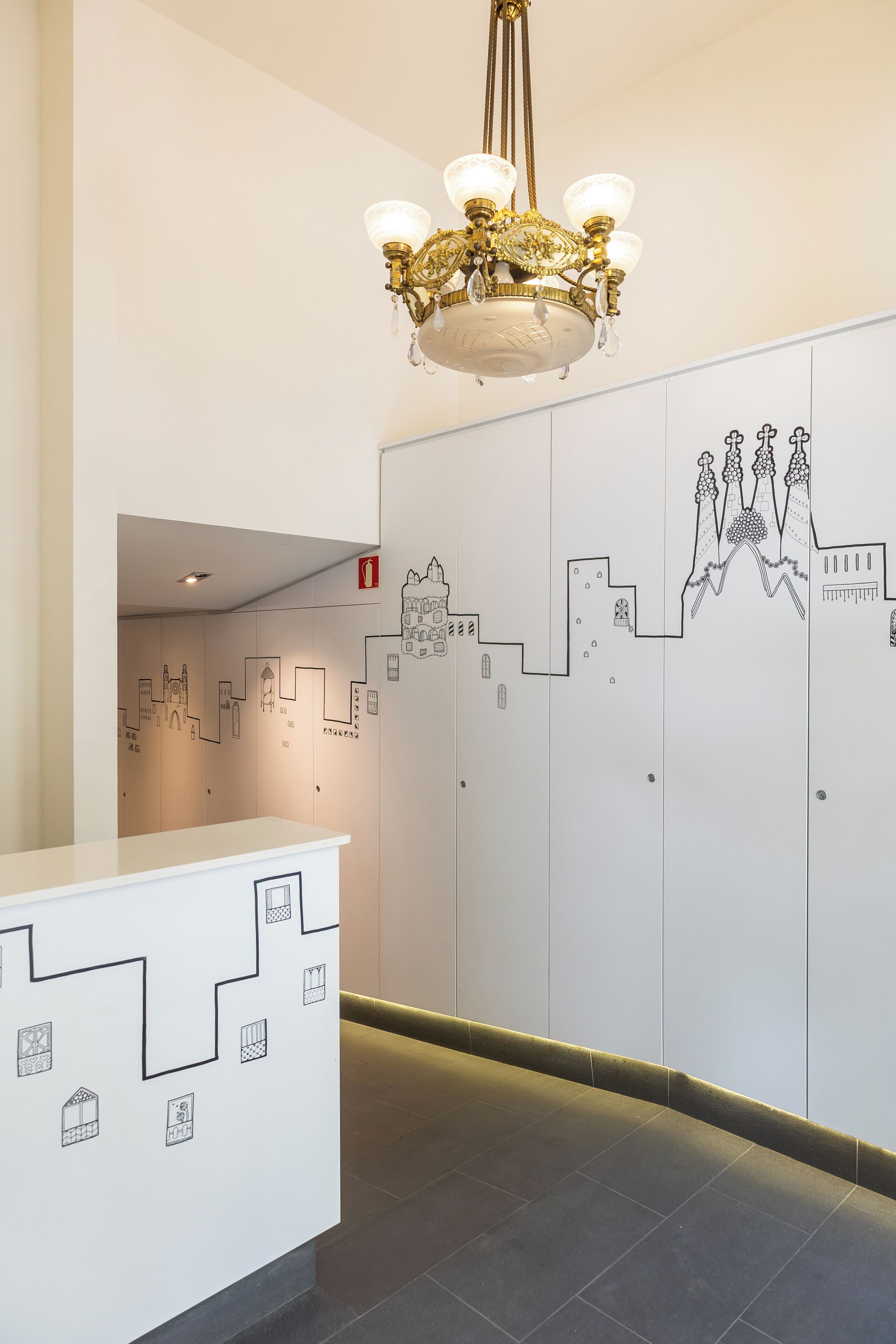 Een hal met een wit receptiebalie, een kroonluchter en een muur met zwart lijntjes tekeningen van gebouwen.