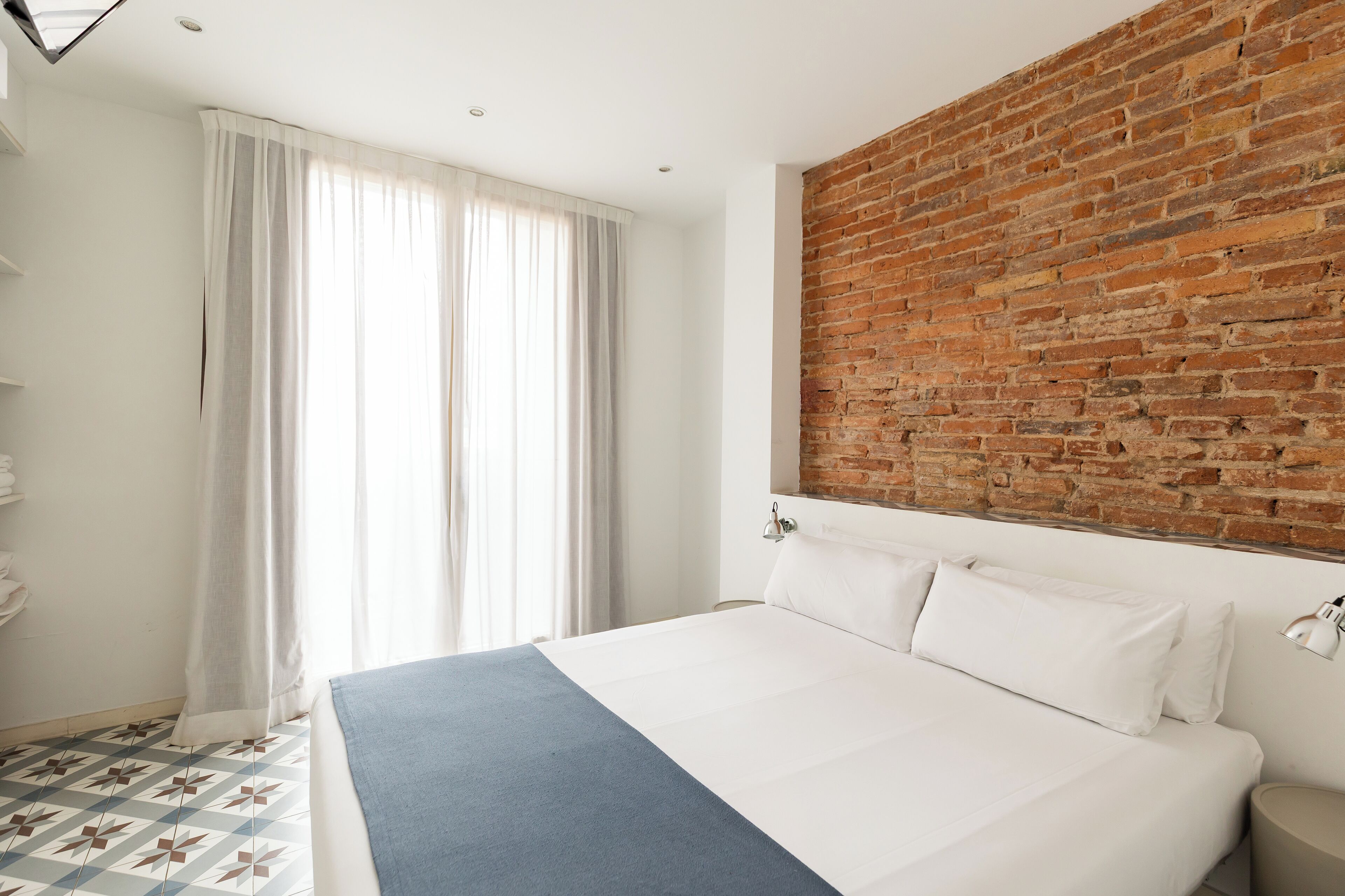 A hotel room with a large bed, a brick accent wall, a window with curtains, and a patterned floor.