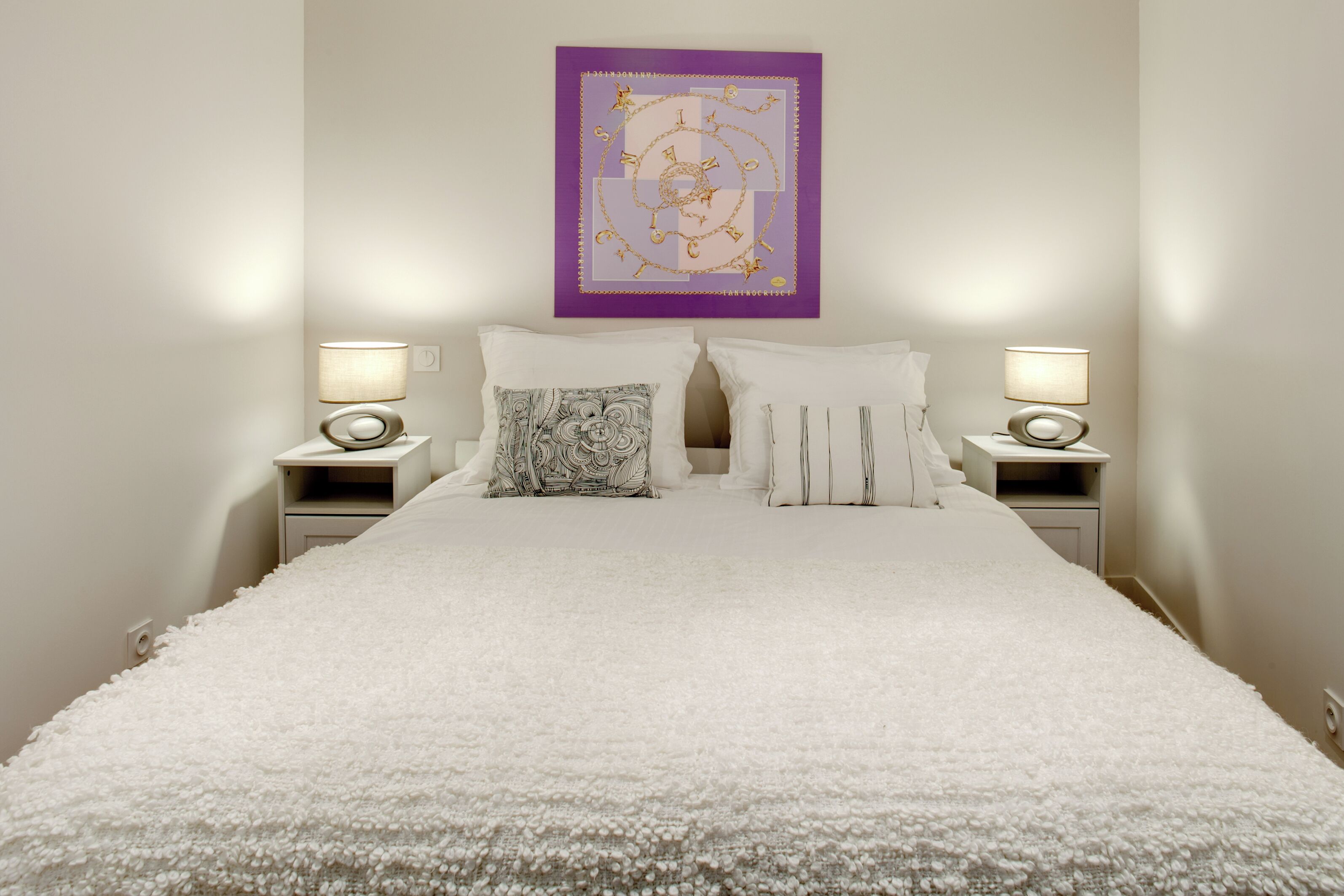 Un letto rifatto con copriletto bianco, due comodini con lampade e un quadro con cornice viola sulla parete.